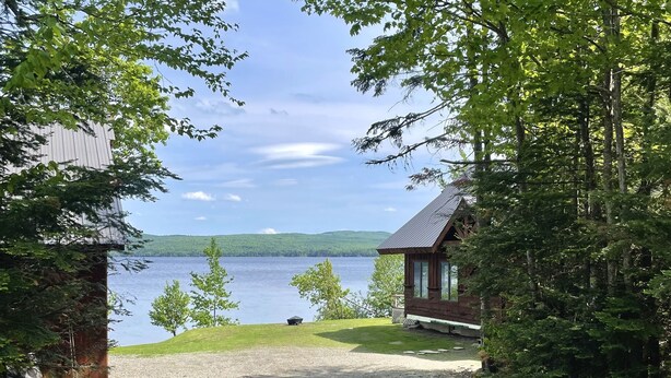 Une allée de gravier mène à un chalet d’où l’on a vue sur un lac et des collines en arrière-plan.