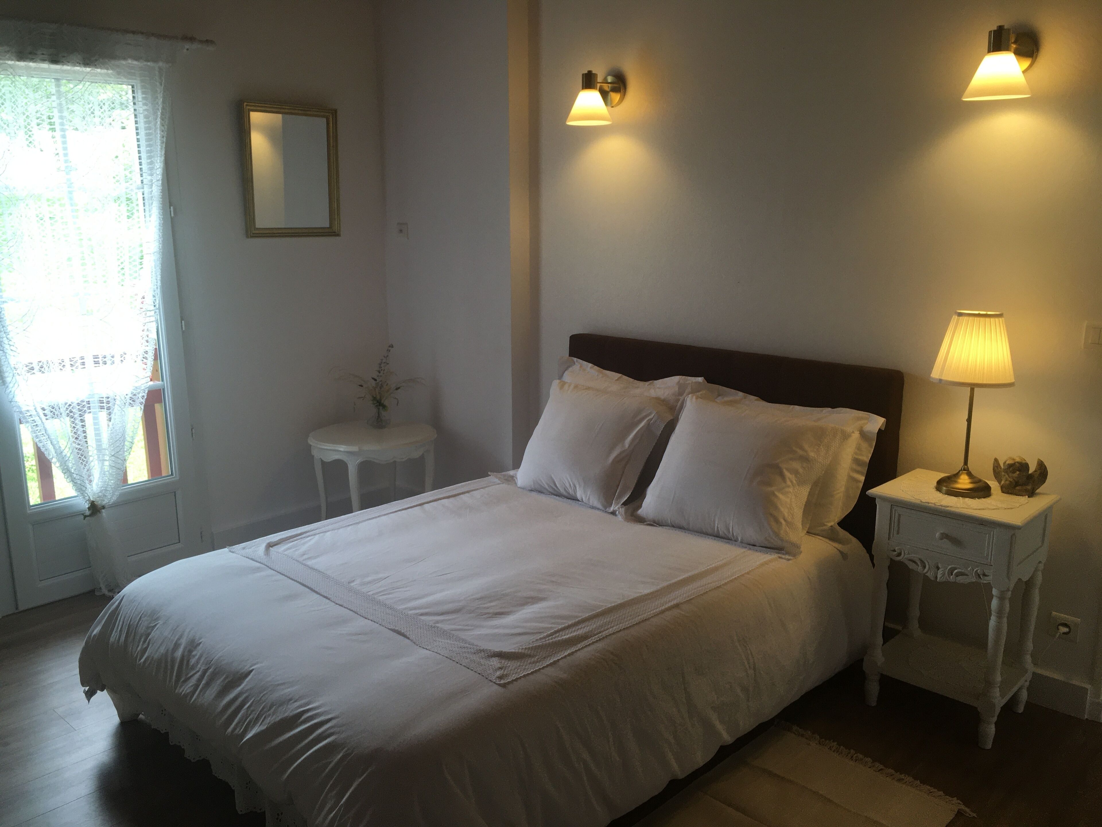 A neatly made bed with white linens, a small bedside table with a lamp, and a wall-mounted light fixture.