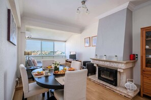 A modern living room with a fireplace, dining area, and a large window offering a city view.