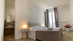 Un dormitorio con cama, lámparas de noche, mesita de noche, baño con espejo y ventana con cortinas.
