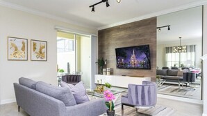 A modern living room with a grey sofa, a coffee table with a book and flowers, a TV on the wall, and a large window. - Amazing Villa 5BR - Private Pool & Themed Rooms - 5 miles Disney! (Kissimmee)