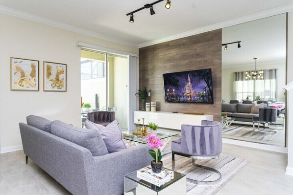 Una sala de estar moderna con un sofá gris, una mesa de centro con un libro y flores, un televisor en la pared y un ventanal grande.