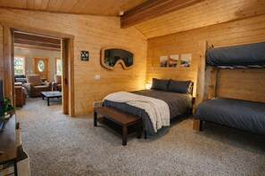 A cozy log cabin room with a bed, bench, and bunk bed, wooden walls, and a view of the outdoors.