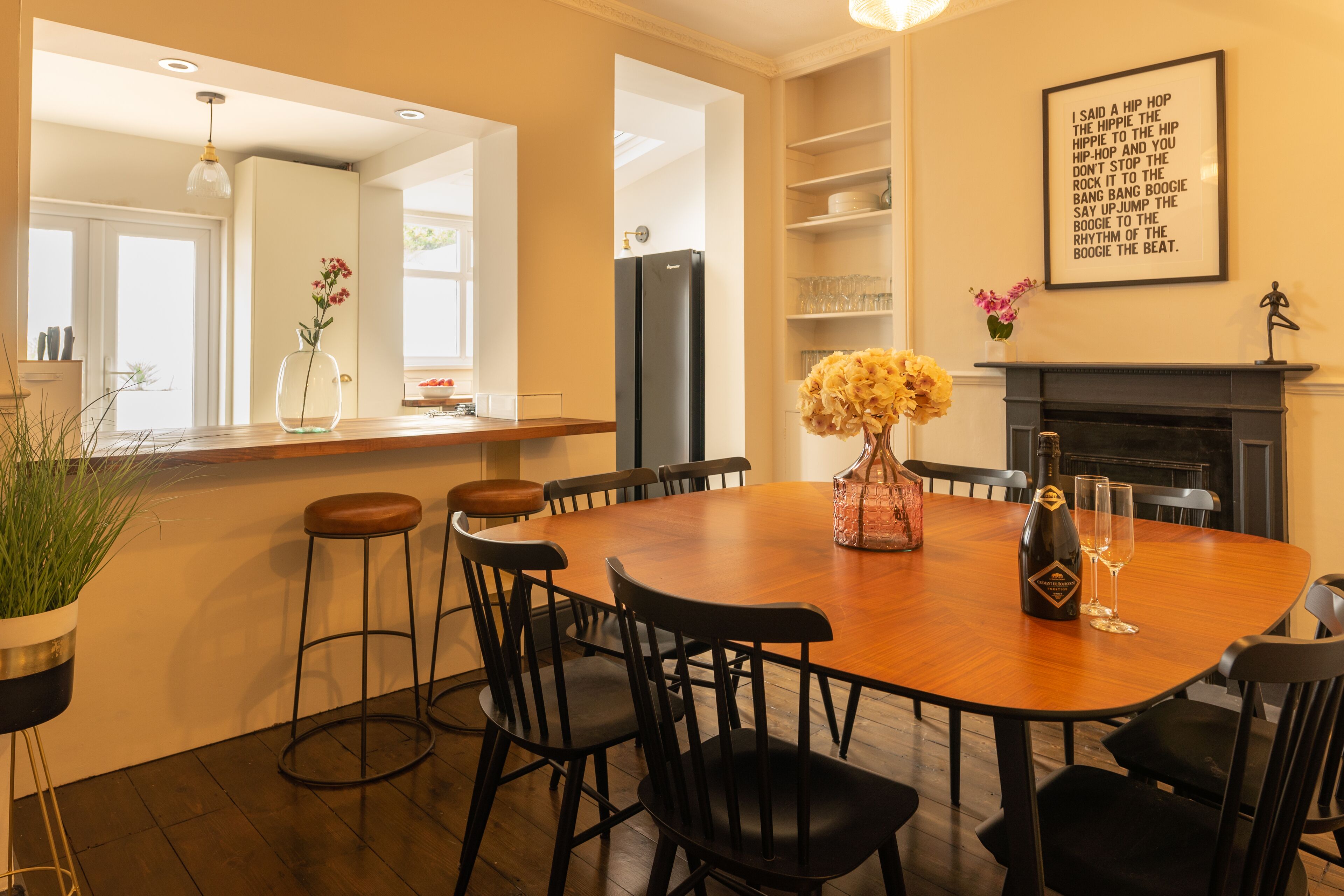 A dining area with a wooden table, black chairs, a vase with flowers, a bottle of champagne, and a framed quote on the wall.