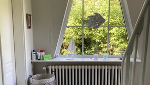 Habitación con ventana que da a zonas verdes, un radiador, una cesta de mimbre y un estante pequeño con objetos.