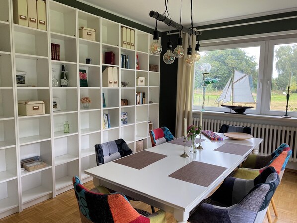 Un comedor con una mesa blanca, sillas de colores, una estantería y vistas al exterior.