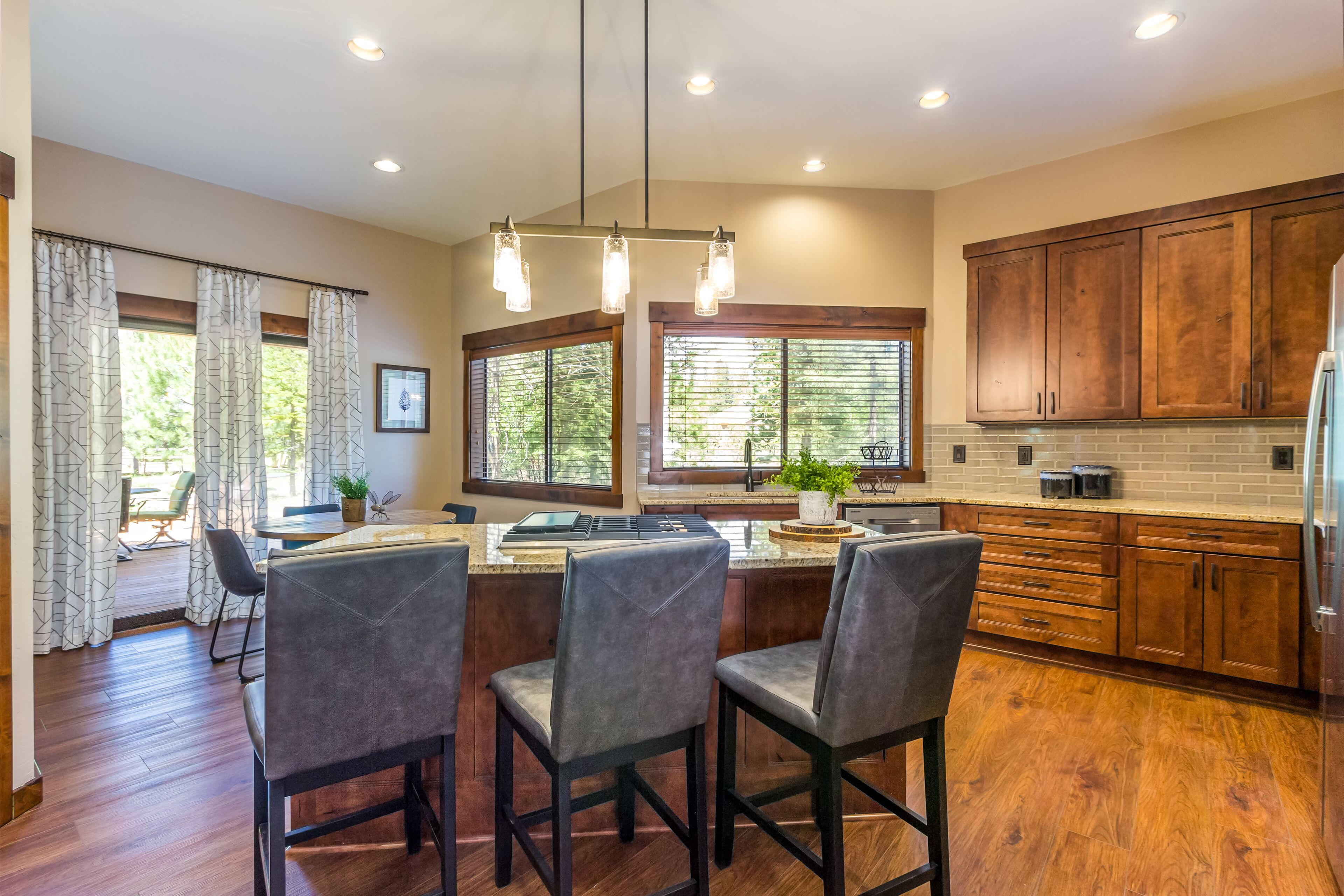 A modern kitchen with wooden cabinets, a central island with bar stools, and large windows providing natural light.