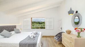 Een slaapkamer met een bed, een raam met uitzicht op groen en bergen, een nachtkastje met een lamp en een kledingkast.