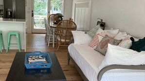 Ein gemütliches Wohnzimmer mit Holzboden, einem schwarzen Couchtisch, einem weißen Sofa mit gemusterten Kissen, einem Essbereich mit Korbstühlen und einer Küche mit Kühlschrank und Schränken.