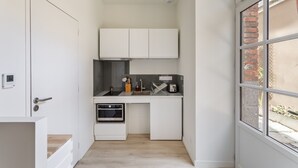 A compact kitchen with white cabinetry, a built-in oven, and a window with a brick wall view. - La Cour des Carmélites 2 - Studio calme en centre-ville (Rennes)