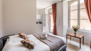 A hotel room with a bed, a small table with a vase, a window with curtains, and a mirror on the wall. - La Cour des Carmélites 2 - Studio calme en centre-ville (Rennes)