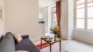 A modern living room with a grey sofa, a small wooden table with a vase and two cups, and a large window with curtains. - La Cour des Carmélites 2 - Studio calme en centre-ville (Rennes)