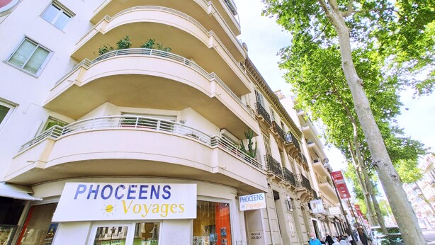 A multi-story building with balconies, a sign for 'PHOCEENS Voyages', and a tree-lined street. - Viva Riviera Palais Holiday Design 2 Bedrooms (Cannes)