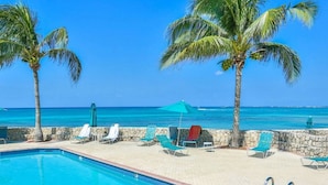 A pool area with lounge chairs and umbrellas, palm trees, and a view of the ocean. - Beach Living at Plantation Village BLGS (Seven Mile Beach)