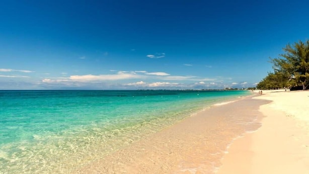 A beach with clear turquoise water, sandy shore, and trees on the right. - Beach Living at Plantation Village BLGS (Seven Mile Beach)