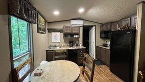 A compact kitchen with a dining table, wooden chairs, a refrigerator, and a window with a view of greenery.