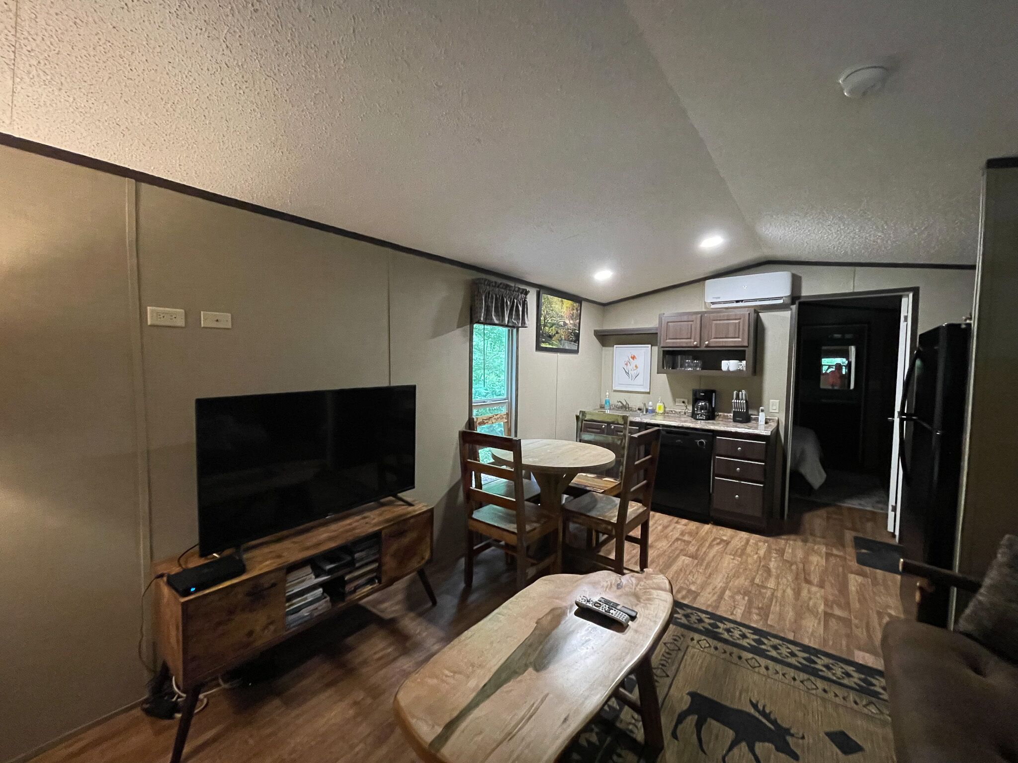 A compact living space with a flat-screen TV, wooden furniture, a dining table, and a kitchen area with cabinets and appliances.