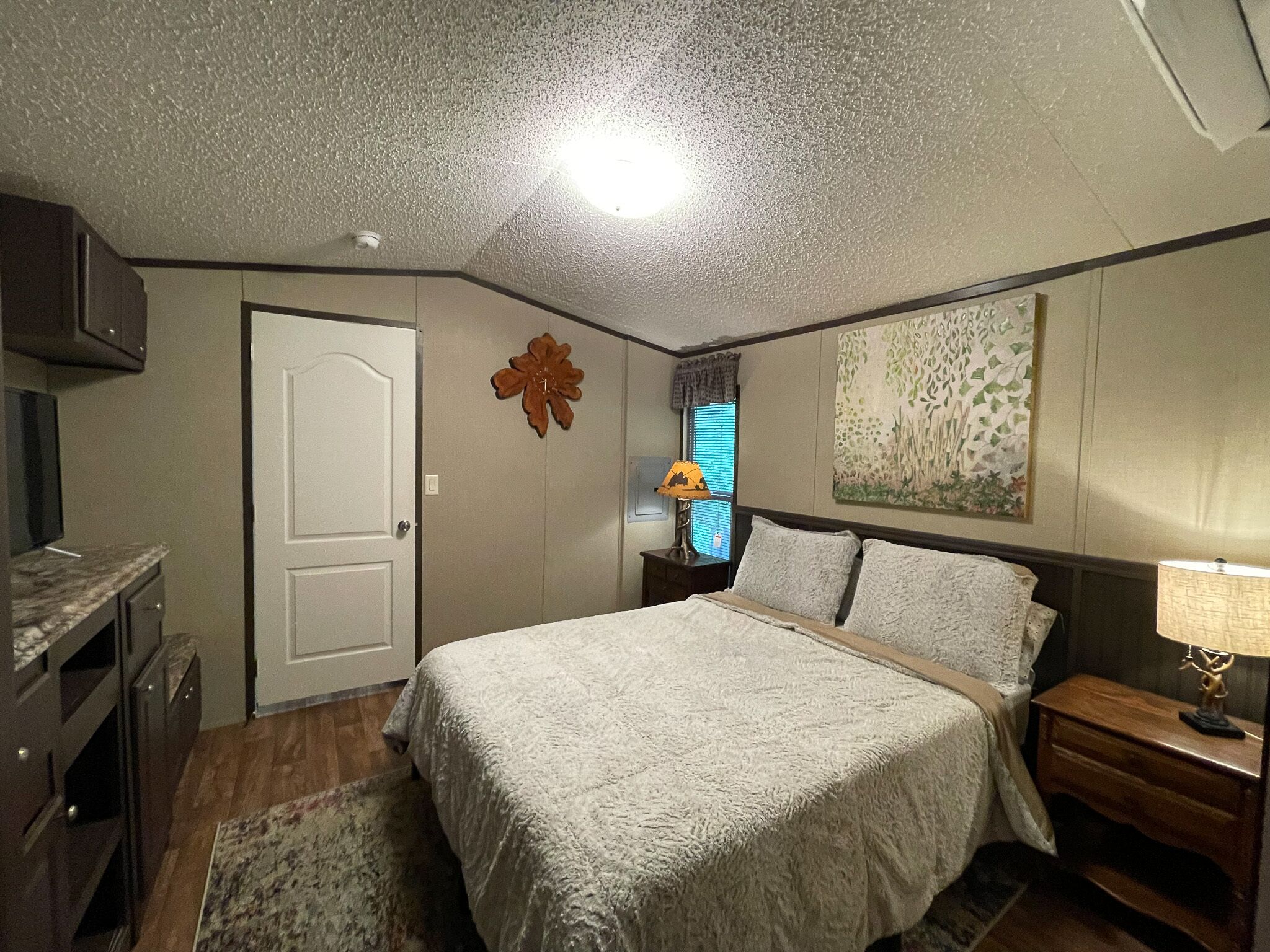 A bedroom with a bed, a nightstand, a lamp, a door, a wall decoration, and a window with blinds.