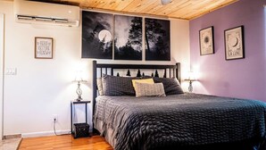 A bedroom with a wooden ceiling, a bed with a dark patterned bedspread, two bedside tables with lamps, and wall art depicting a moon and trees. - Moonlight Cottage - Romantic Smoky Mountain Cabin with Hot Tub & Community Pool (Sevierville)