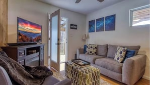 A living room with a grey sofa, a television, a patterned ottoman, and a ceiling fan. - Moonlight Cottage - Romantic Smoky Mountain Cabin with Hot Tub & Community Pool (Sevierville)