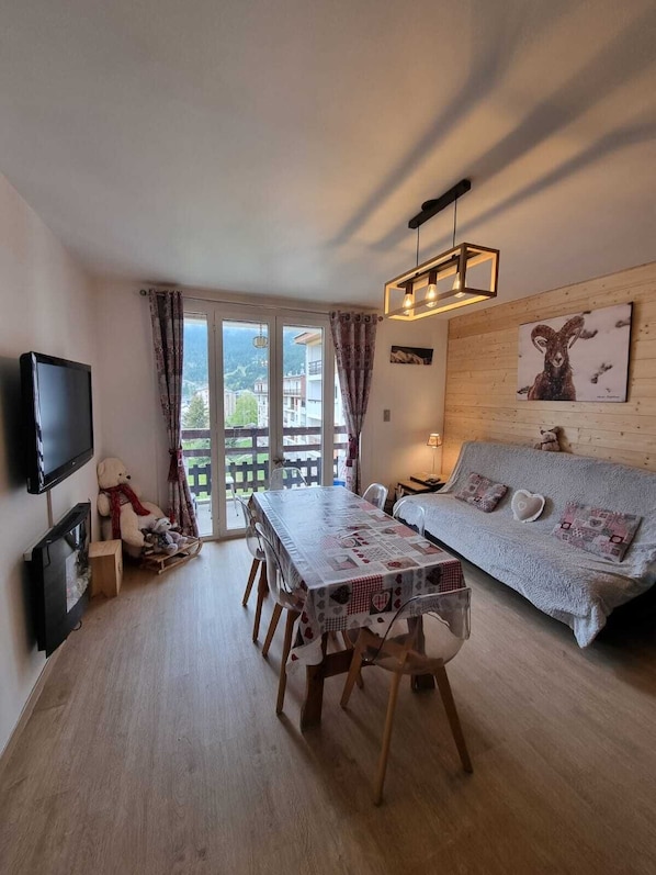 A cozy living room with a wooden floor, a dining table with chairs, a sofa, a TV, and a large window with curtains.