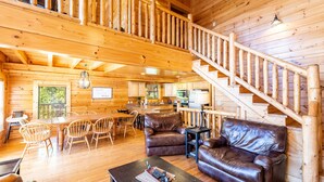 Interior de una cabaña de madera con comedor, dos sofás de cuero, cocina y una escalera de madera.