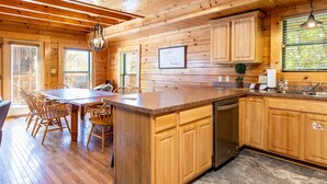 Interior de una cabaña de madera con comedor, cocina y un amplio ventanal con persianas.
