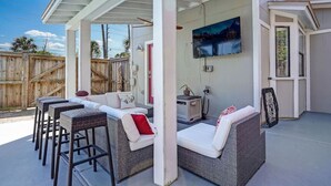 Une terrasse extérieure couverte, aménagée avec des meubles en osier, un téléviseur fixé au mur et une clôture en bois en arrière-plan.