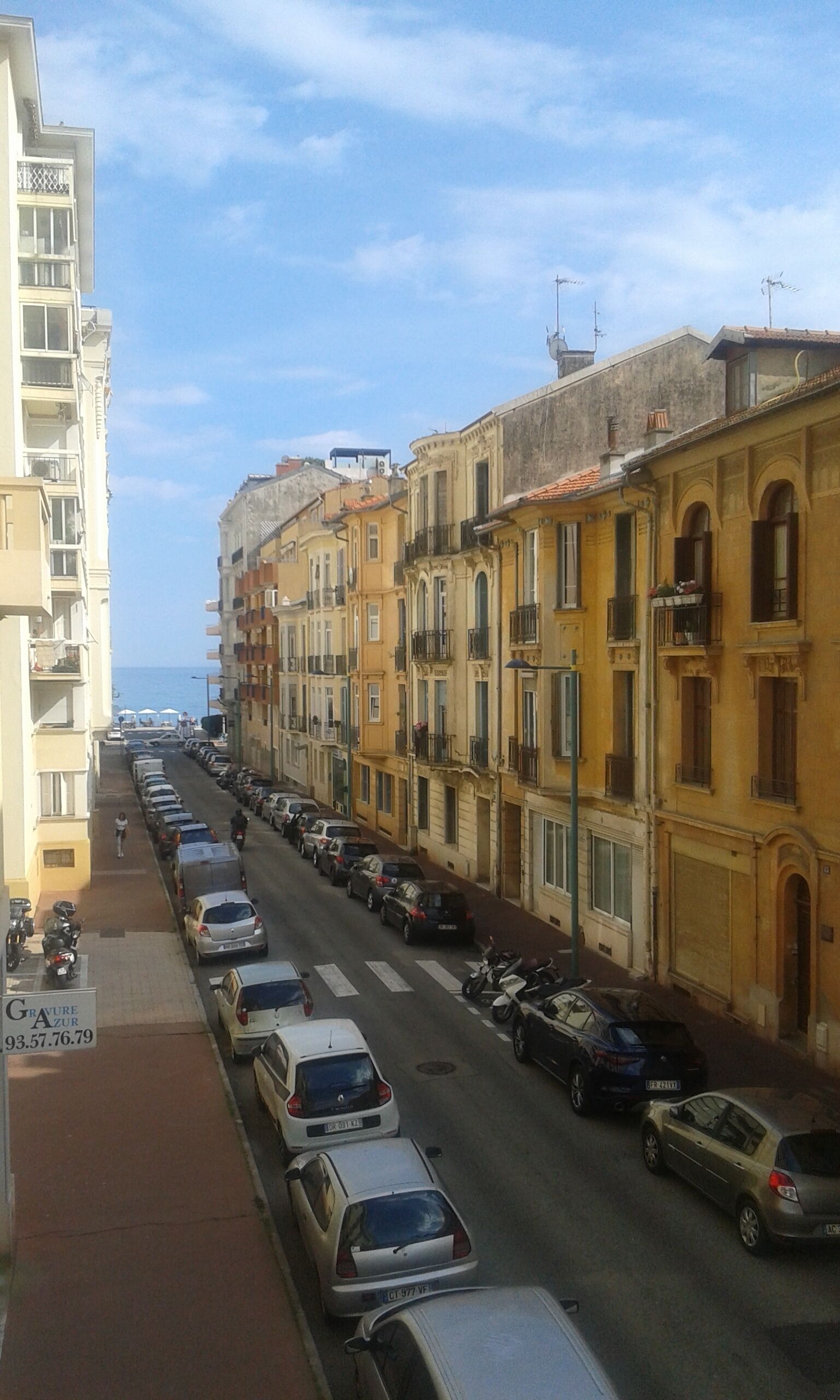 A street lined with multi-story buildings, parked cars, and a clear sky.