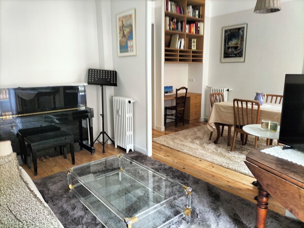 Uma sala de estar com um piano, uma mesa de vidro, uma estante de livros e um espaço de jantar com mesa e cadeiras.