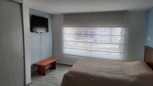 Un dormitorio con cama, una banca, un televisor montado en la pared y una ventana con vista a edificios.