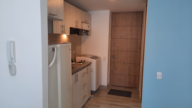 Cocina compacta con refrigerador, microondas y estufa. Se observa una puerta de madera y un interruptor de luz en la pared.