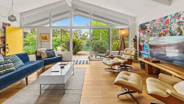 A spacious living room with a large TV, comfortable seating, and a view of greenery through large windows.