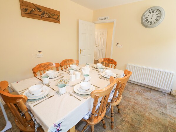 A dining room with a table set for four, wooden chairs, a clock on the wall, and a door in the background.