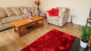 Ein Wohnzimmer mit Couchtisch aus Holz, zwei gemusterten Sofas, einem roten Teppich, einer Wanduhr, einer Topfpflanze und einem kleinen Beistelltisch.