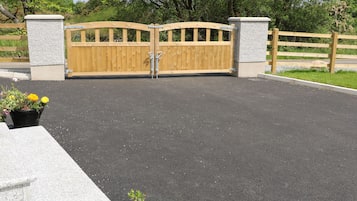 Una entrada con reja de madera y pilares de piedra, rodeada por un área pavimentada y vegetación.