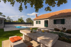 En stenbelagt terrasse med et bord og stole, en swimmingpool samt en have med blomster og planter.