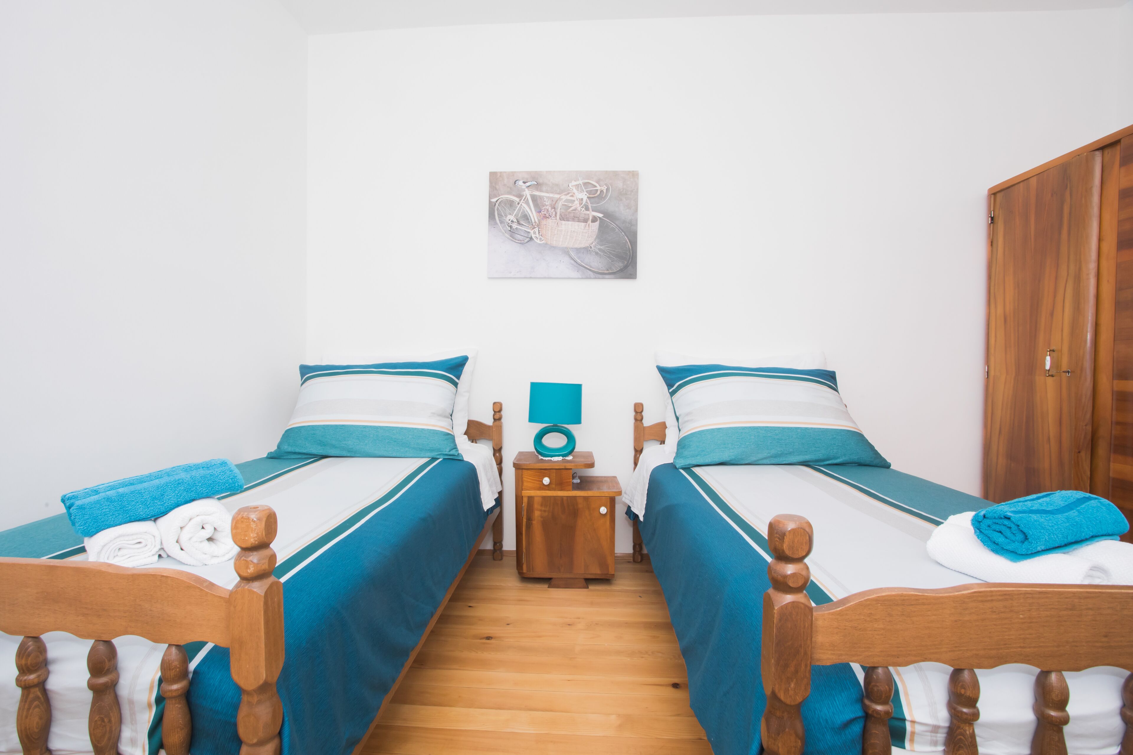 Una habitación con dos camas individuales, cabeceros de madera, una mesita de noche de madera con una lámpara y un cuadro colgado en la pared de una bicicleta.