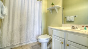 Un baño con inodoro blanco, cortina de ducha, lavamanos con espejo y un estante pequeño con una planta.
