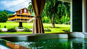 Une vue depuis une fenêtre, à travers un rideau, donnant sur une maison sur pilotis et une piscine.