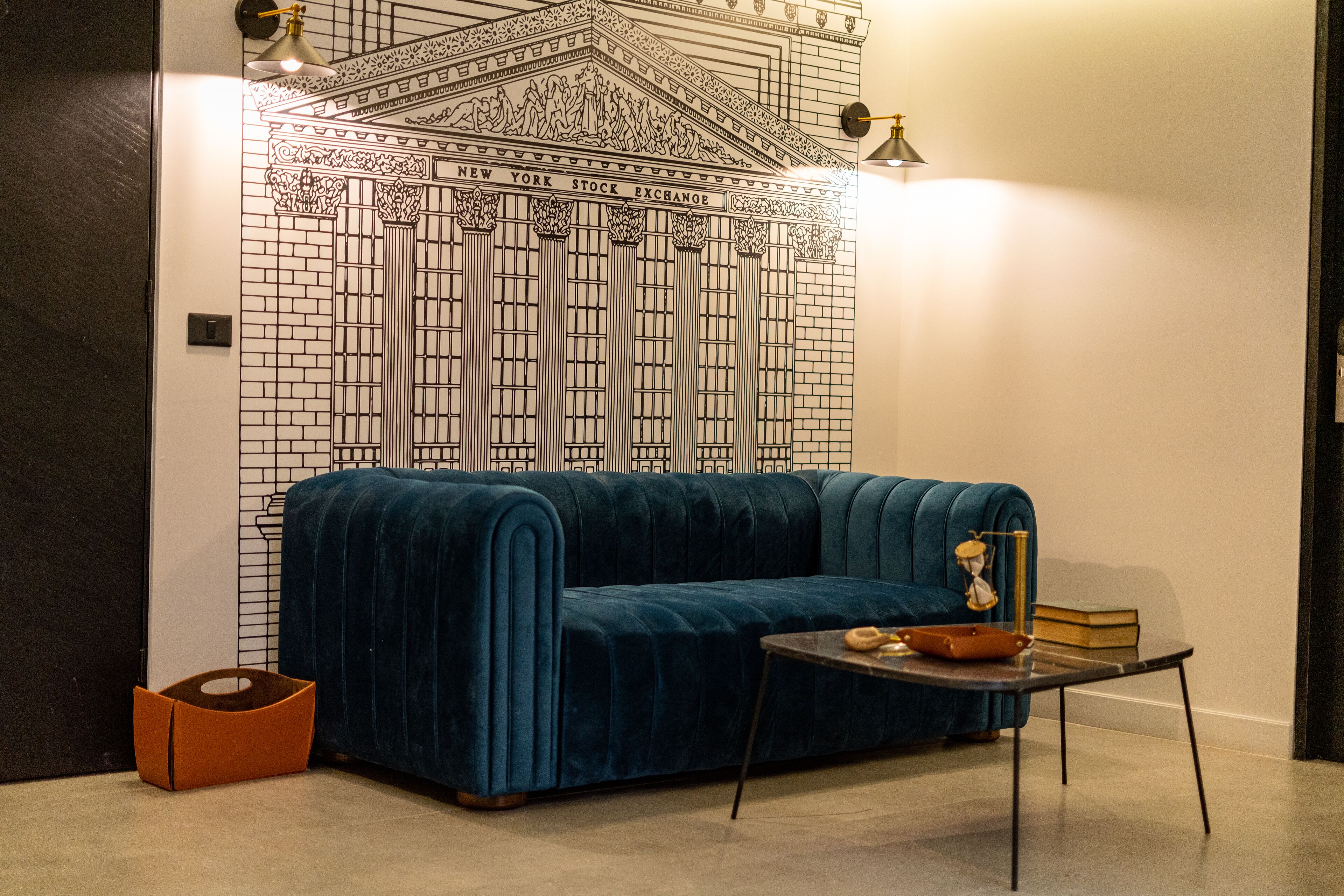 A room with a blue velvet sofa, a small table with books and a lamp, and a wall mural of the New York Stock Exchange.
