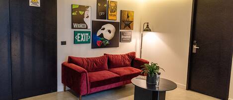 A red velvet sofa, a small round table with a plant, and wall art including a cat, a lion, and a Broadway sign.