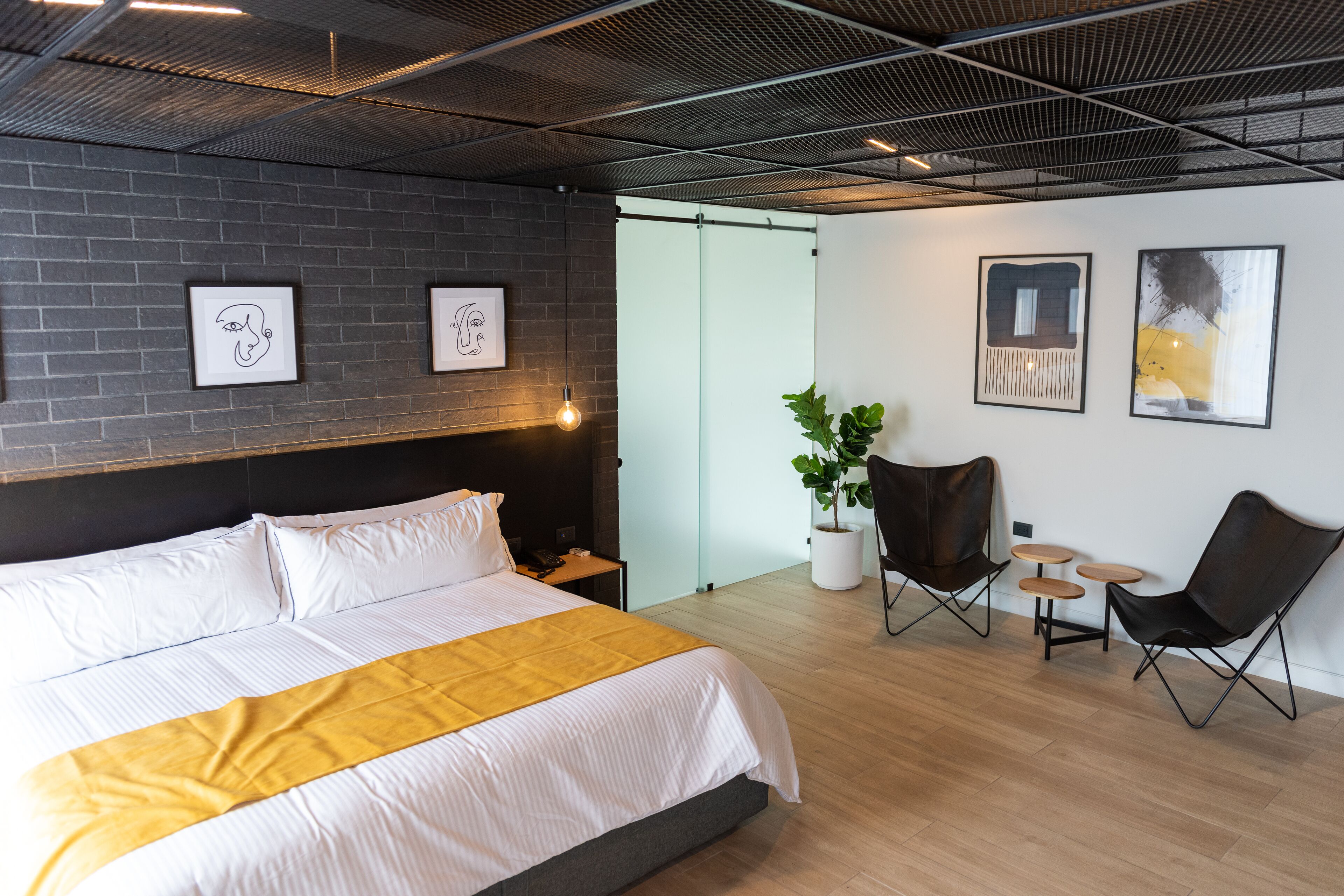 A modern hotel room with a large bed, a small desk, a potted plant, and two chairs.
