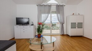 Ein modernes Wohnzimmer mit Glastisch, Flachbildfernseher auf einem weißen Schrank, einer Topfpflanze und Blick durch eine Glastür.