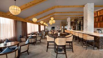 Une salle à manger spacieuse avec des poutres en bois, des tables rondes, des chaises en osier et des luminaires sphériques suspendus.