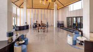 Eine großzügige Lobby mit modernem Design, inklusive einer zentralen Sitzecke mit runden Tischen und blauen Stühlen, einer Rezeption und einem Kronleuchter.