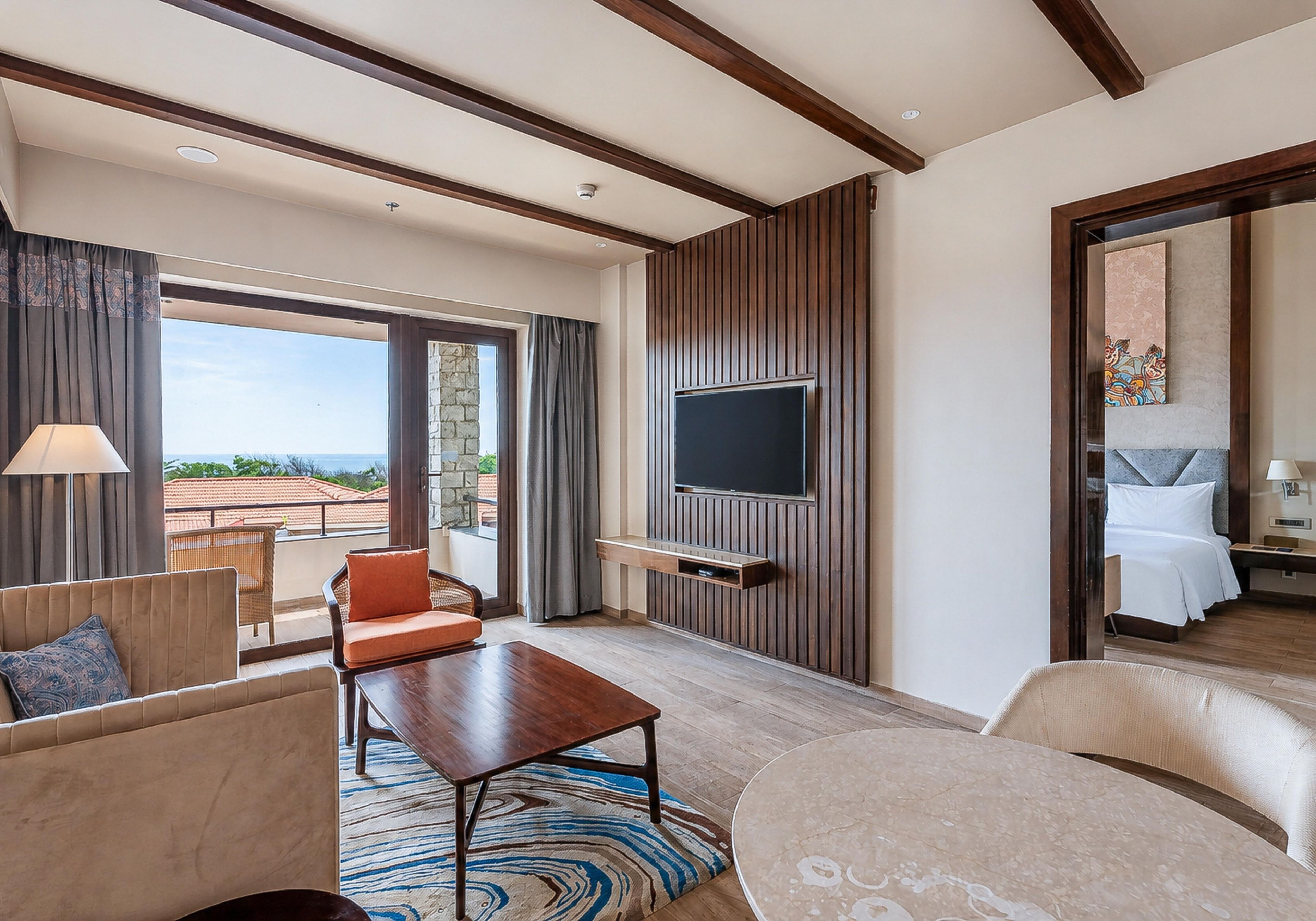 A hotel room with a large TV mounted on a wooden panel wall, a balcony with a view, and a bed with a white duvet.