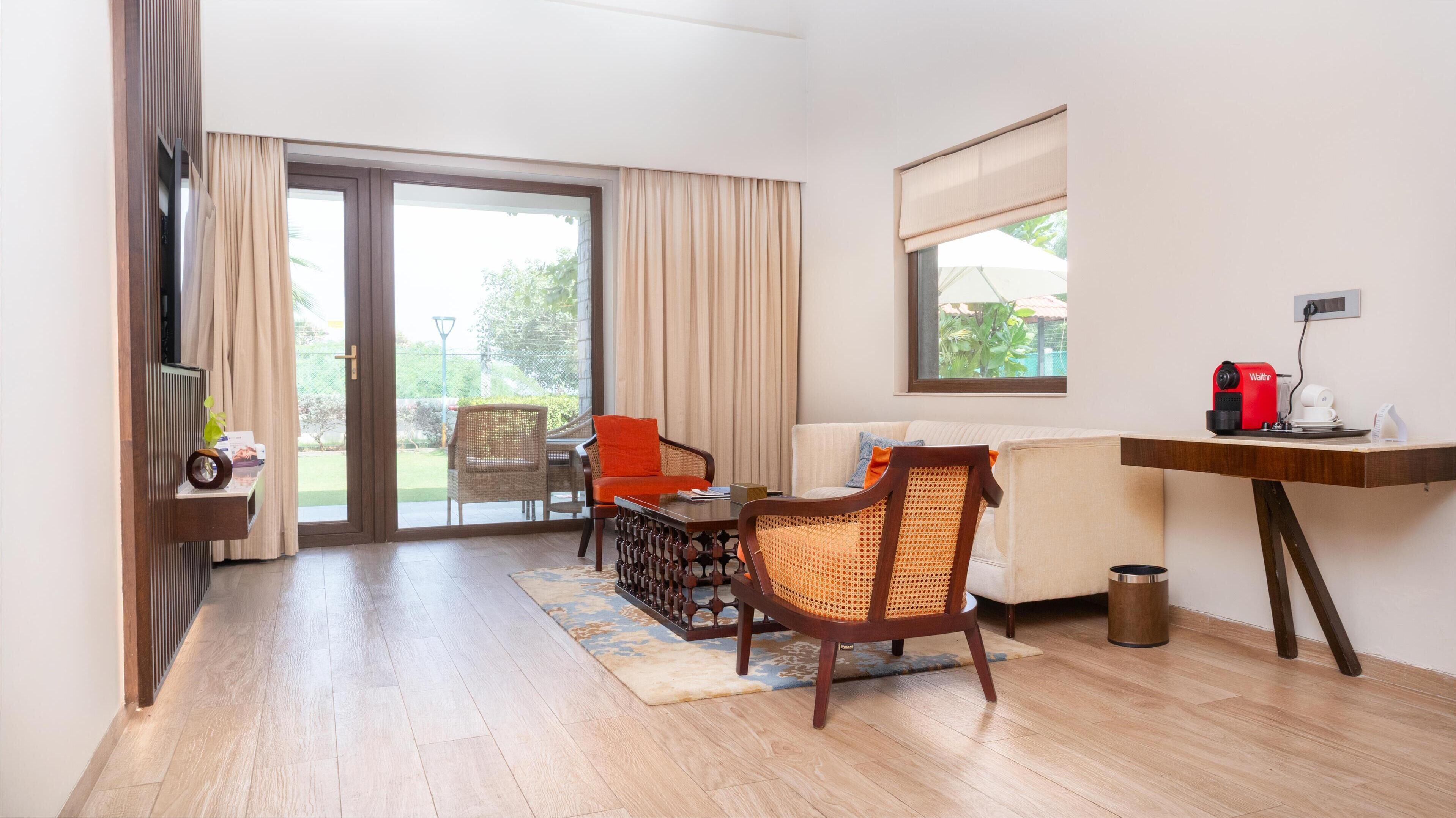 A hotel room with a sliding glass door to a balcony, a sofa, a chair, a small table, and a desk with a coffee machine.