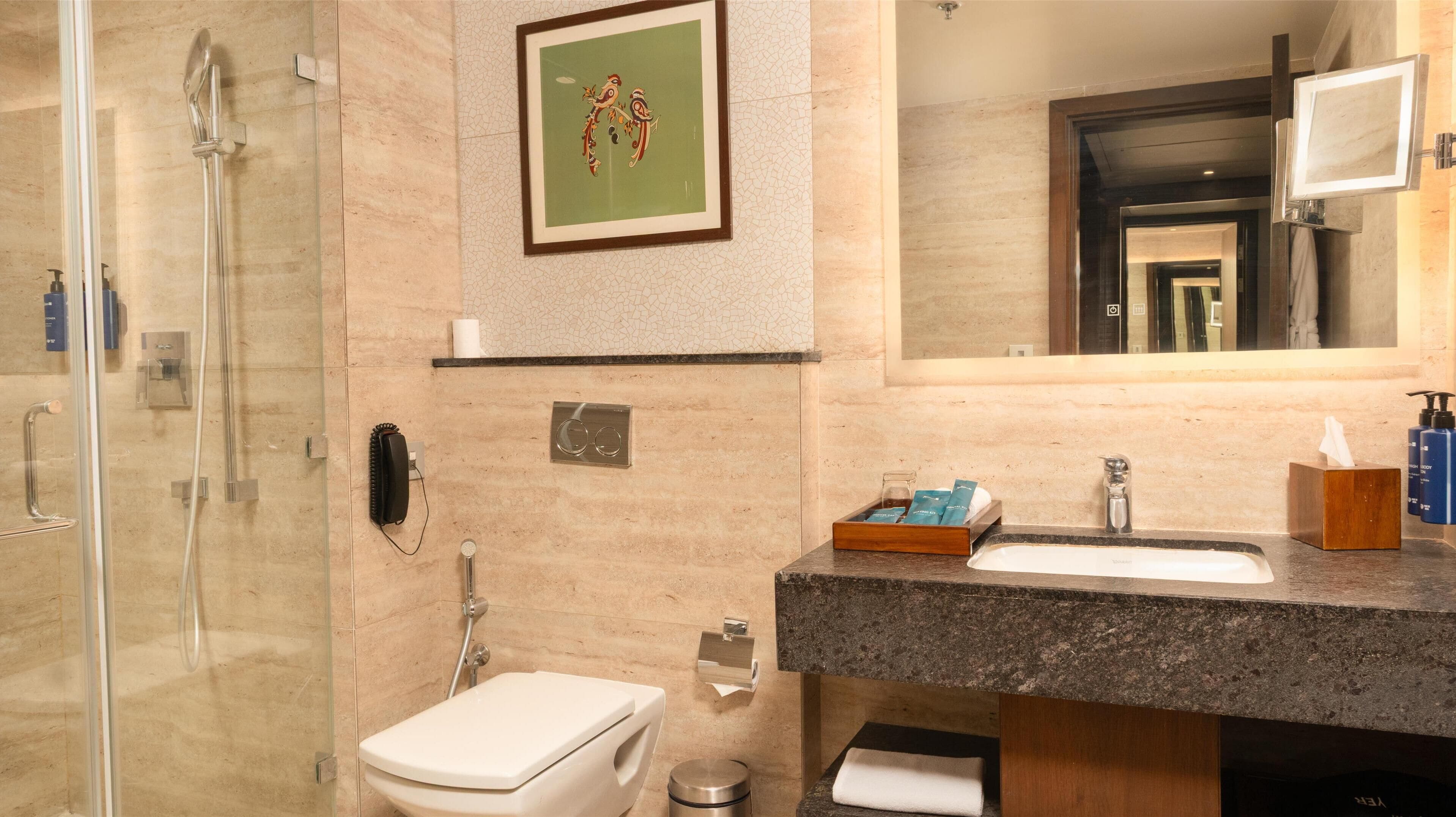 A modern bathroom with a glass shower enclosure, a toilet, a sink with a marble countertop, and a framed artwork above the mirror.