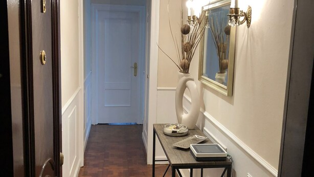 Un pasillo con piso de madera, una puerta azul, una pared blanca, un candelabro y una mesa con un florero y un espejo.
